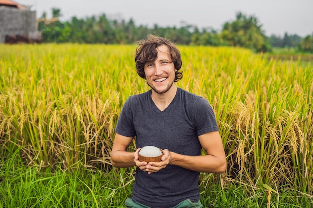 Man is holding a cup of boiled rice in a wooden cup on the background of a ripe rice field