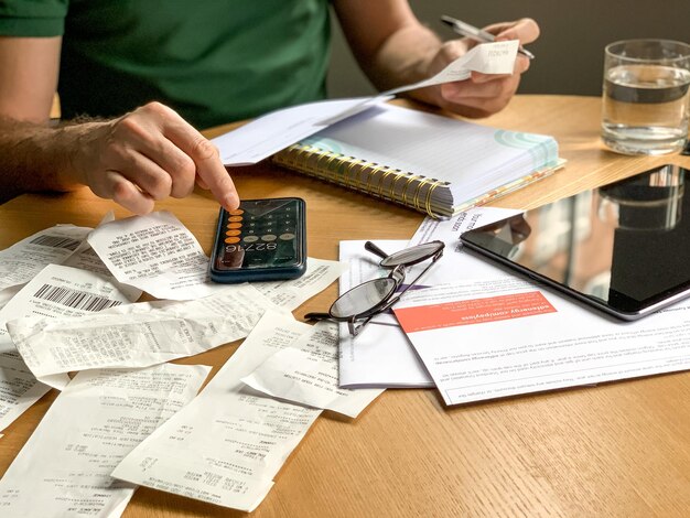 Photo man is making audit of household expenses using calculator and notebook lots of receipts and bills