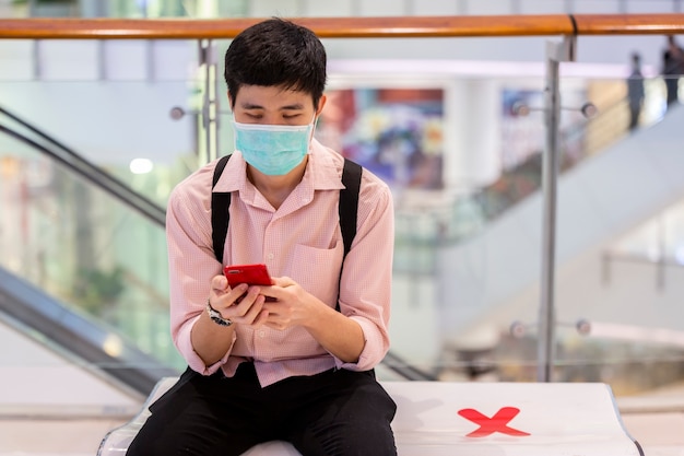 Un uomo sta giocando uno smartphone mentre è seduto su una panchina del centro commerciale pubblico con cautela segnali sociali di allontanamento. (messa a fuoco selettiva)