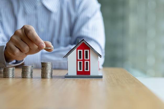 A man is putting coins on a pile piles of coins on a table and a house model on it saving money to spend and managing to buy a future home Money saving ideas to buy a house or residence