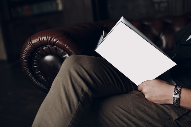 The man is reading book with blank white cover on leather brown chair