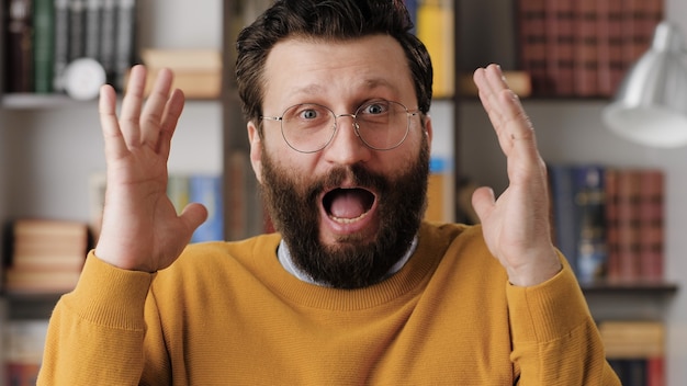 Man is scared, terrified. Frightened bearded man in glasses in office or room in apartment with suddenly throws his hands up and looking at camera, he is very scared and just terrified. Close-up view