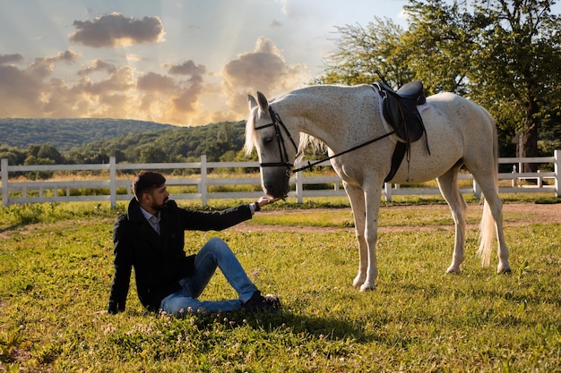 Man is sitting on the grass and feeding a horse