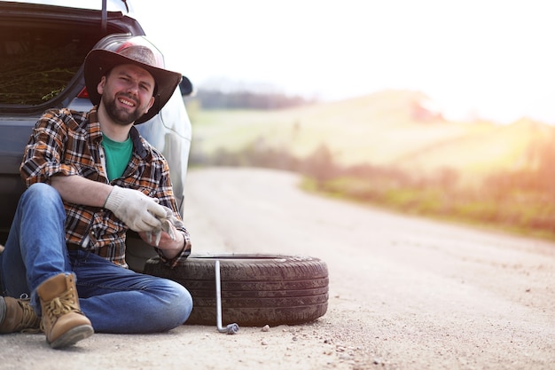 The man is sitting on the road by the car in the nature