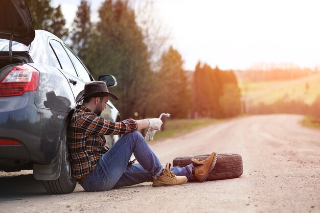 The man is sitting on the road by the car in the nature