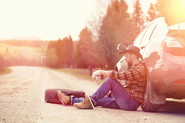 The man is sitting on the road by the car in the nature