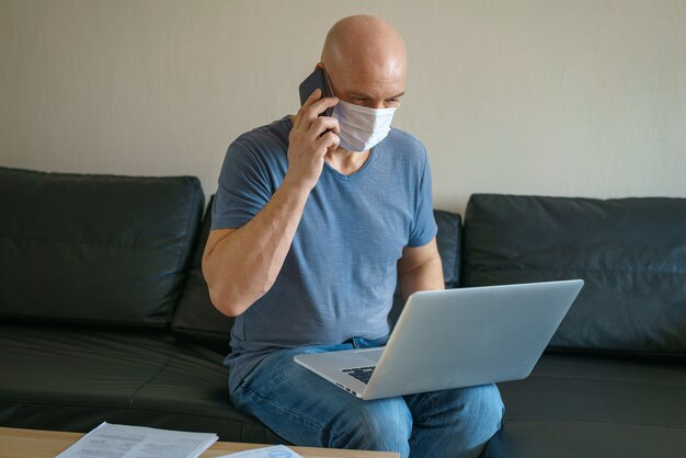 Man is sitting on a sofa in a protective mask with a laptop and phone, remote work in quarantine