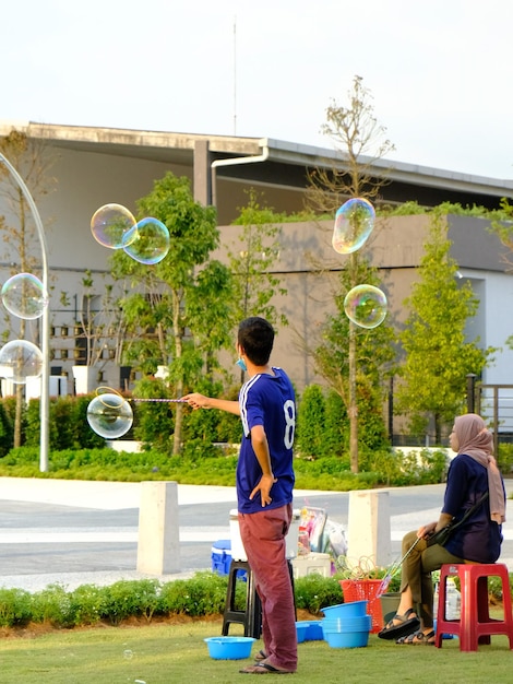 A man is sitting on a stool and watching bubbles in front of a building.