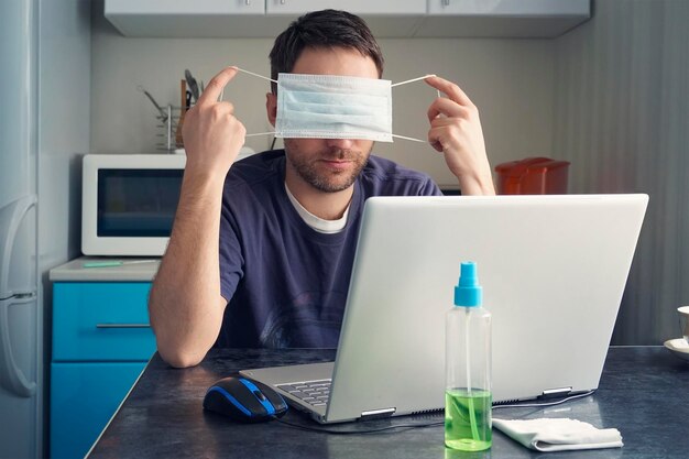 Un uomo è seduto a un tavolo e lavora su un laptop in abiti domestici sullo sfondo della cucina. stanco del lavoro a distanza e della quarantena, il ragazzo si toglie la maschera protettiva dal viso.