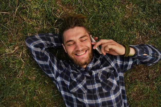 Man is talking on phone using iPhone 12 mini Young Caucasian man in shirt with dreadlocks and beard lies in clearing in park and smiles View from above Moscow Russia May 7 2022