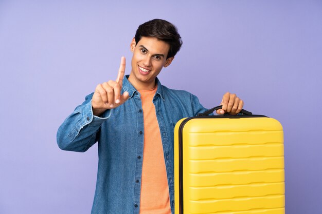 Man over isolated purple wall in vacation with travel suitcase and counting one