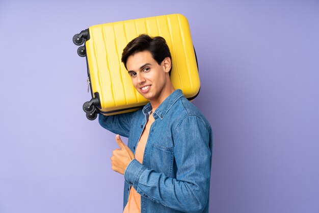 Man over isolated purple wall in vacation with travel suitcase and with thumb up