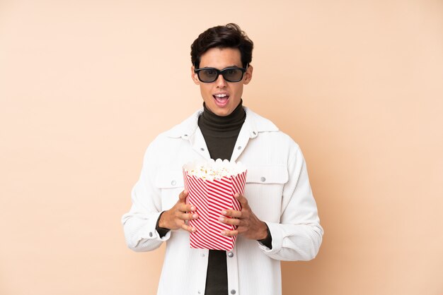 Man over isolated wall surprised with 3d glasses and holding a big bucket of popcorns