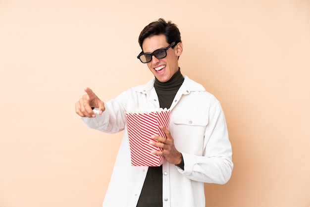 Man over isolated wall with 3d glasses and holding a big bucket of popcorns while pointing away