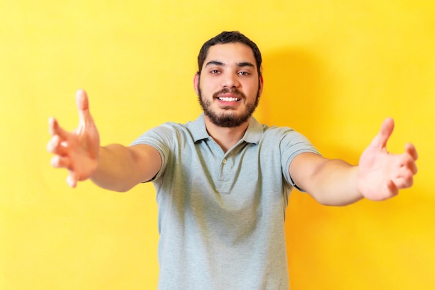 Man over isolated yellow background looking at the camera smiling with open arms for hug Cheerful expression embracing