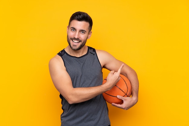 Man over isolated yellow wall playing basketball and pointing to the lateral