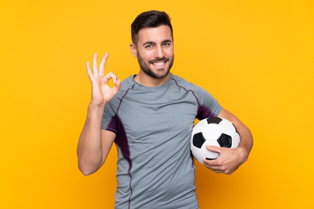 Man over isolated yellow wall with soccer ball and making OK sign