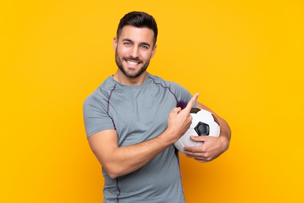 Uomo sopra la parete gialla isolata con pallone da calcio e che punta verso il lato