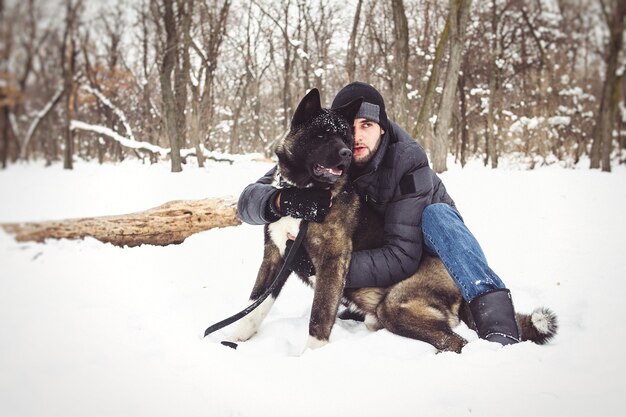 A man in a jacket and a knitted hat walks through a snowy forest with an American Akita dog