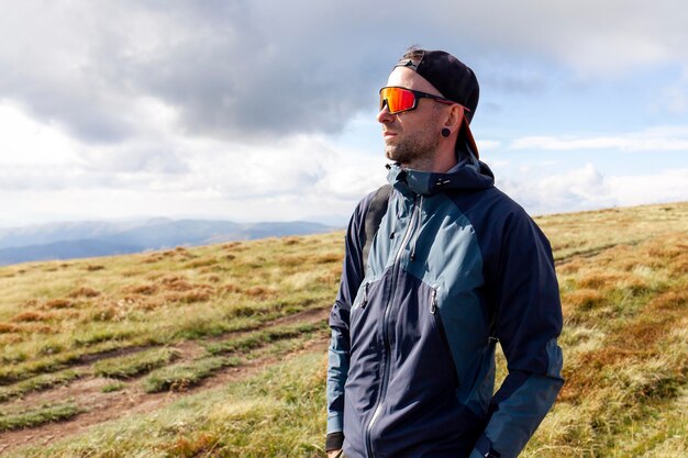 man in jacket and sunglasses stands on the mountain and looks at the sun tourist guy in mountain