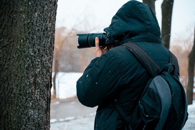 a man in a jacket with a hood and a briefcase on his back, holding a camera and taking pictures 