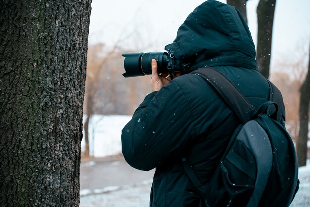 A man in a jacket with a hood and a briefcase on his back, holding a camera and taking pictures 