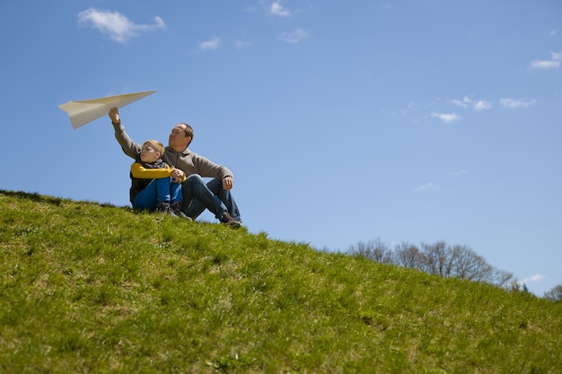 Man and kid boy sitting on top of hill and going to start paper airplane Father and son together