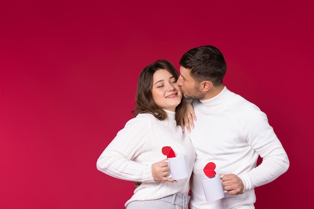 A man kisses on the cheek a smiling woman on a red background. White cups for tea and lovers' time.