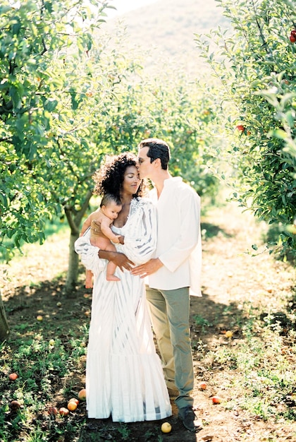 Man kisses on the forehead a woman with a newborn in her arms in an apple orchard