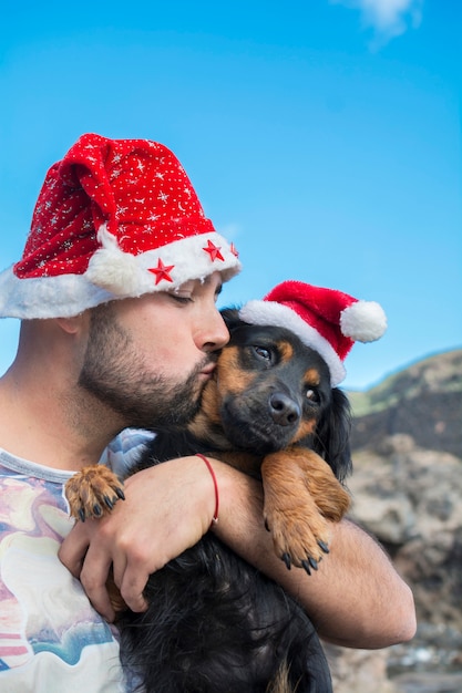 クリスマス帽子をかぶってクリスマスのために彼の犬にキスをする男性