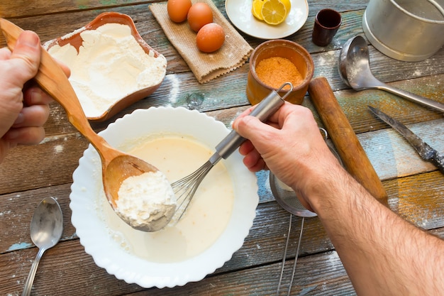 The man knead the dough with a whisk. Wheat flour, batter, eggs, a lemon and kitchen utensils 