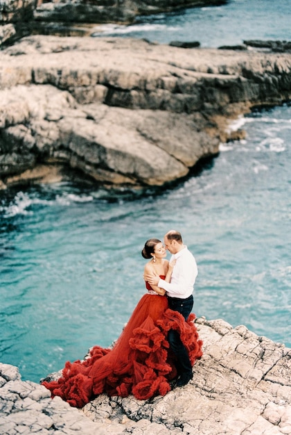 Man knuffelt een vrouw in een rode jurk terwijl hij op de stenen boven de zee staat