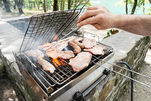 Man koken vlees op barbecue Chef-kok grillen van vlees in park buiten in de buurt van meer Concept van het eten van bbq buiten met gasgrill in de zomer Focus op de hand van de man Warm filter met terug zonlicht