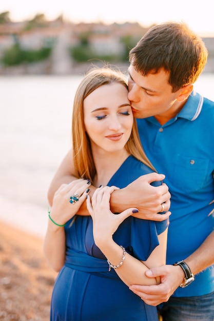 Man kust zachtjes lachende zwangere vrouw in een blauwe jurk knuffelen haar schouders