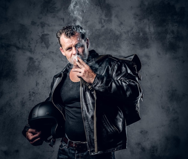 Un uomo in giacca di pelle tiene il casco da motociclista e fuma una sigaretta.