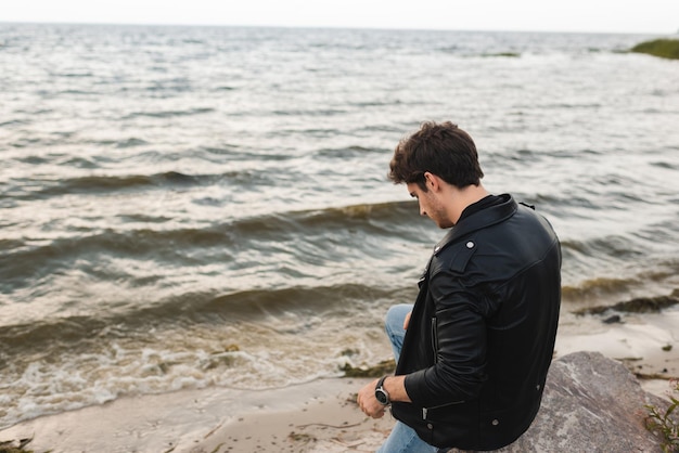 Man in leather jacket sitting on stone on sea coast