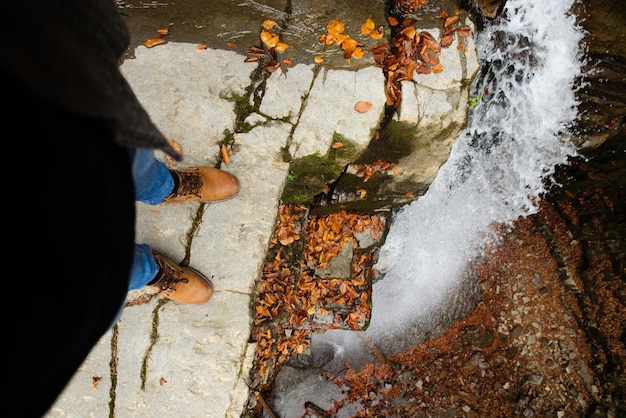 Gambe dell'uomo in blue jeans e stivali marroni seduti sul bordo guardando la cascata