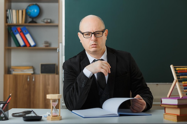 Man leraar met bril controleren klasse register kijken camera met sceptische uitdrukking loensende ogen zittend op school bureau voor schoolbord in klas
