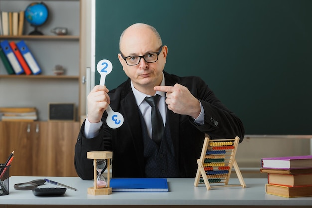 Man leraar met bril zittend aan school bureau met klassenregister voor bord in klaslokaal met nummerplaten die les uitleggen met sceptische uitdrukking