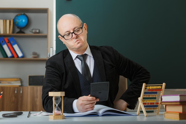 Man leraar met een bril die aan de schoolbank zit met klassenregister voor het bord in de klas en uitleg geeft over de les die naar de rekenmachine kijkt