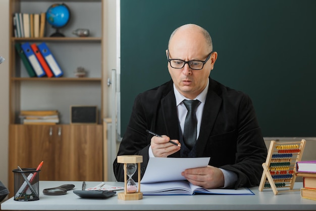 Man-leraar met een bril die aan de schoolbank zit voor het schoolbord in de klas en huiswerk controleert van studenten die verrast kijken