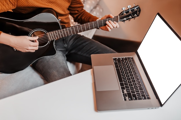 Man leren gitaar spelen met behulp van online leren thuis. Guy zittend aan tafel met laptop en zwarte gitaar