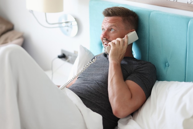 Photo man lies in bed in hotel room and talk on phone.