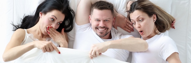 Man liggend met twee geliefden in bed vrouwen die onder dekking kijken en zich afvragen promiscue seks