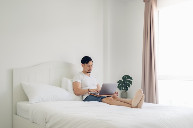 Man liggend op bed werken op zijn laptop in gezellige witte slaapkamer.