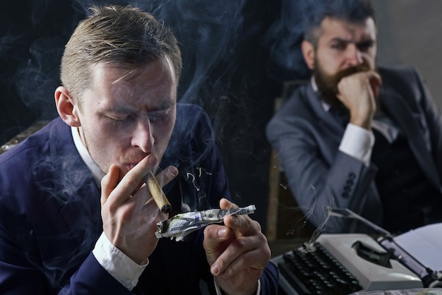 Man lighting cigar from burning banknote businessman smoking cigar at business meeting squander concept businessmen in suits sit at table with typewriter and money in dark interior defocused