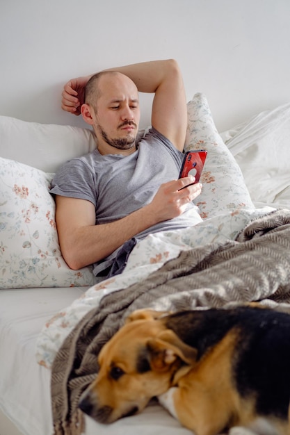 Man ligt op het bed met hond die ontspant en telefoon gebruikt om video te kijken en nieuws te lezen terwijl hij op mobiel werkt