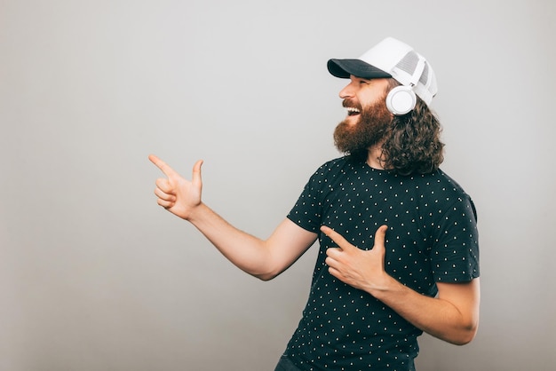 Man listening music on his new headphones is pointing at a free space over gray background wearing a black shirt and a white cap