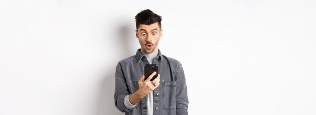 Man look surprised at smartphone screen receive exciting news on phone standing against white backgr