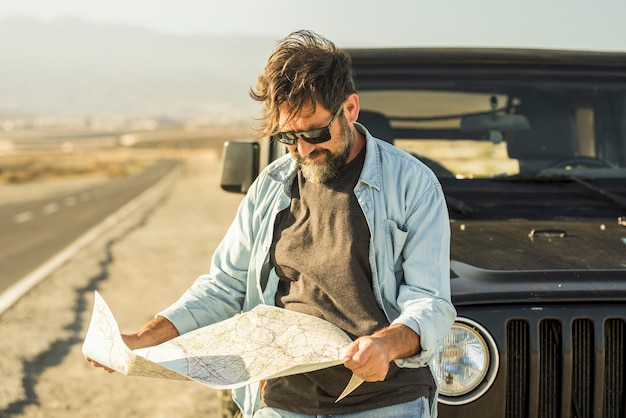 道端で車に寄りかかって地図を見ている男。車の外に立っている紙の地図で目的地の場所を確認する成熟した男。道端で紙の地図を使って航路を探している男